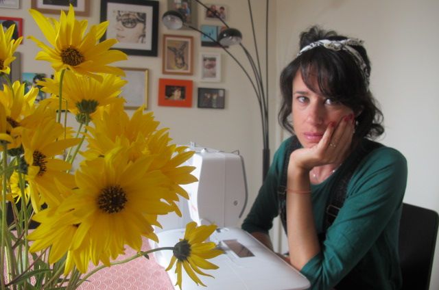 Elena OsesB, una chica morena de pelo corto con una cinta en la cabeza, está sentada en una silla y apoya sus brazos sobre una mesa. A su vez apoya su barbilla sobre su mano izquierda. Delante de ella hay una máquina de coser blanca y, al lado, un gran ramo de margaritas amarillas