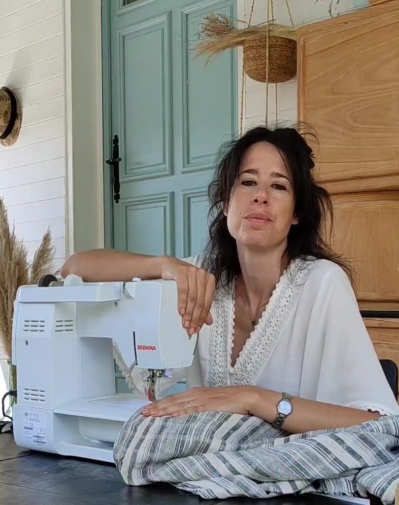 Elena Oses B, una mujer de pelo moreno y con una blusa blanca mira hacia la cámara. Está sentada junto a una mesa sobre la que hay una máquina de coser blanca. Su mano derecha está apoyada sobre la parte superior de la máquina. Su mano izquierda reposa sobre la tela, de rayas grises y blancas, que se encuentra en la máquina.