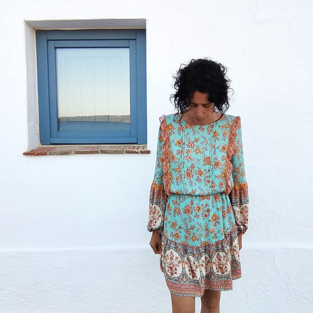Elena Oses, una mujer alta, esbelta, de cabello oscuro y rizado se encuentra de pie, mirando hacia el suelo. Lleva un vestido estampado de manga larga con jaretas y falda fruncida. Arriba, a la izquierda, hay una ventana pequeña, cuadrada y de madera, en color azul.