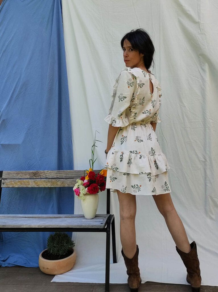 Elena OsesB, una mujer de cabello moreno, recogida, alta y estilizada posa frente a la cámara. Está de pie y de espaldas, pero gira su torso hacia la izquierda para mirar a la cámara. Lleva un vestido corto en color crudo con motivos de bouquets de flores en tonos verdes, naranjas y blancos. Lleva puestas unas botas marrones tipo cowboy. A su izquierda se encuentra un banco de madera sobre el que se encuentra un jarrón blanco con un ramo de flores rojas, blancas, rosas y naranjas. De fondo hay dos sábanas colgadas, a la izquierda una azul y a la derecha una blanca.