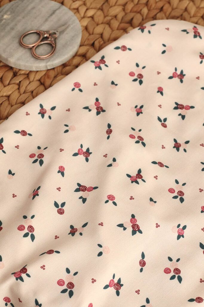 Una tela de color blanco crema, con estampado de ramilletes de flores pequeñas en tonos rojos y rosas con hojas verdes, se encuentra sobre un mantel de esparto. Arriba, a la izquierda de la tela, se encuentra un posavasos de mármol sobre el que reposa una pequeña tijera corta hilos.