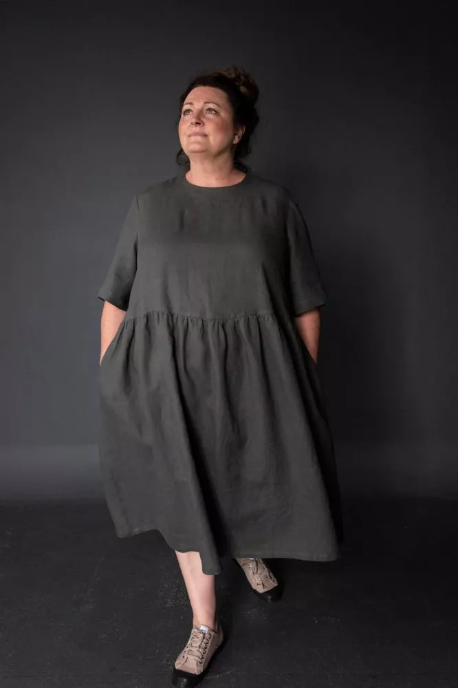 Mujer de mediana edad, con pelo recogido en un moño. Tiene pose de andar y mira hacia arriba. Lleva un vestido ancho, de color gris oscuro, de manga corta y con frunces en la cintura.