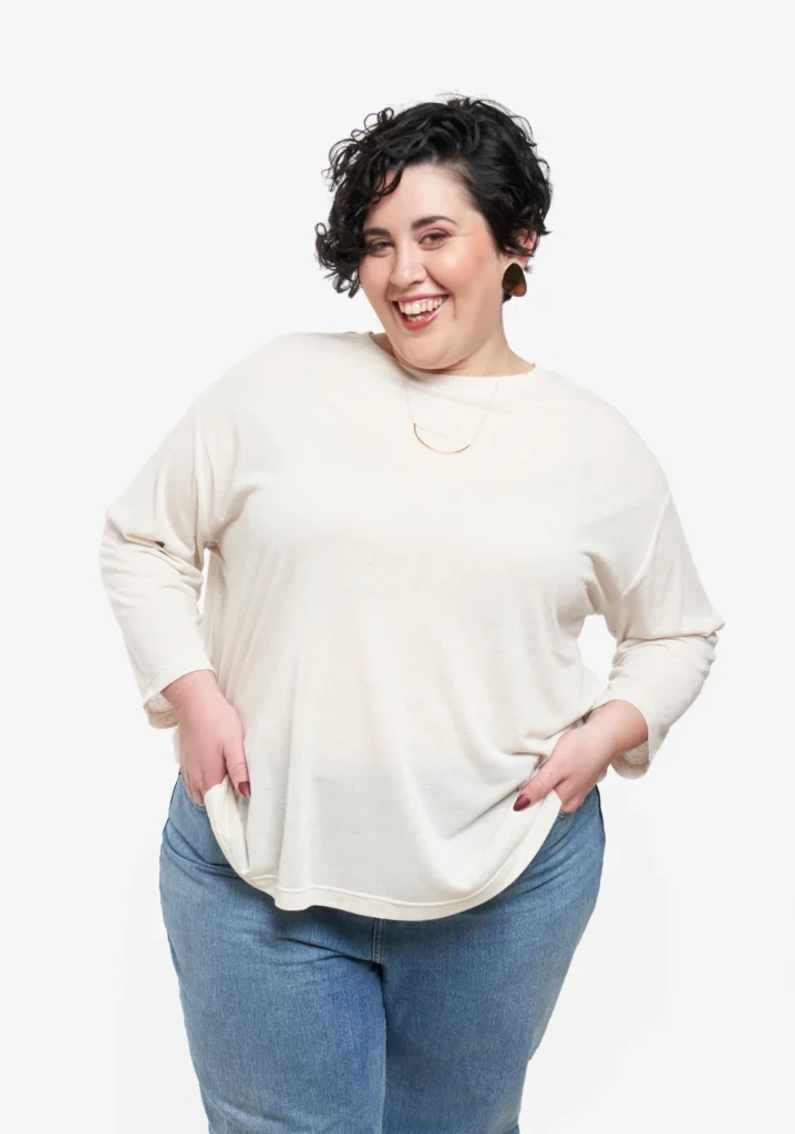 Mujer de pelo corto y rizado con expresión sonriente lleva una camiseta blanca de manga larga y mete las manos en los bolsillos de sus pantalones vaqueros.