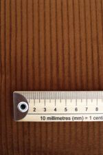 Plano cercano de una tela de pana marrón. Encima de ella se encuentra una regla de madera que indica el tamaño de los bordones, aproximadamente de 5 mm cada uno.
