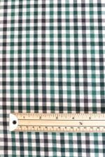 Tela de sarga de algodón de cuadros en distintos tono de verde y crudo. Sobre ella hay una regla de madera, mostrando así el tamaño del cuadro, que es aproximadamente de medio centímetro.
