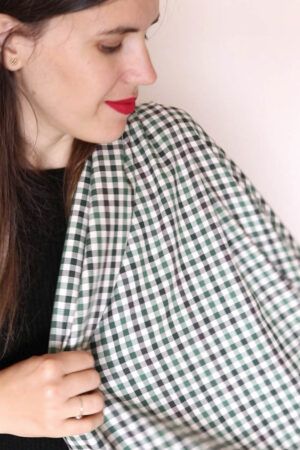 Una chica vestida de negro y con pelo largo, castaño sostiene sobre su hombro, mientras la mira, una tela de cuadros en distintos tono de verde y crudo.