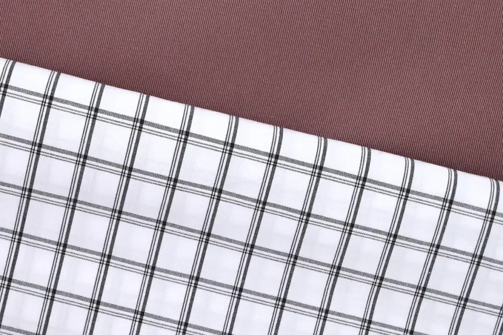 Dos telas deadstock, una de las alternativas al algodón convencional, una encima de la otra. Una es una sarga rosa y otra una tela blanca con raya negras verticales y horizontales, que forman cuadros
