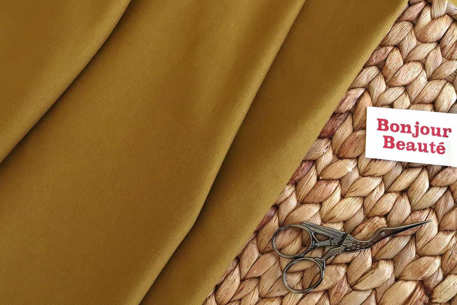 Tela de punto jersey de algodón orgánico en color oliva junto a unas pequeñas. tijeras plateadas con forma de pájaro y una etiqueta de tela en la que pone "Bonjour Beauté"