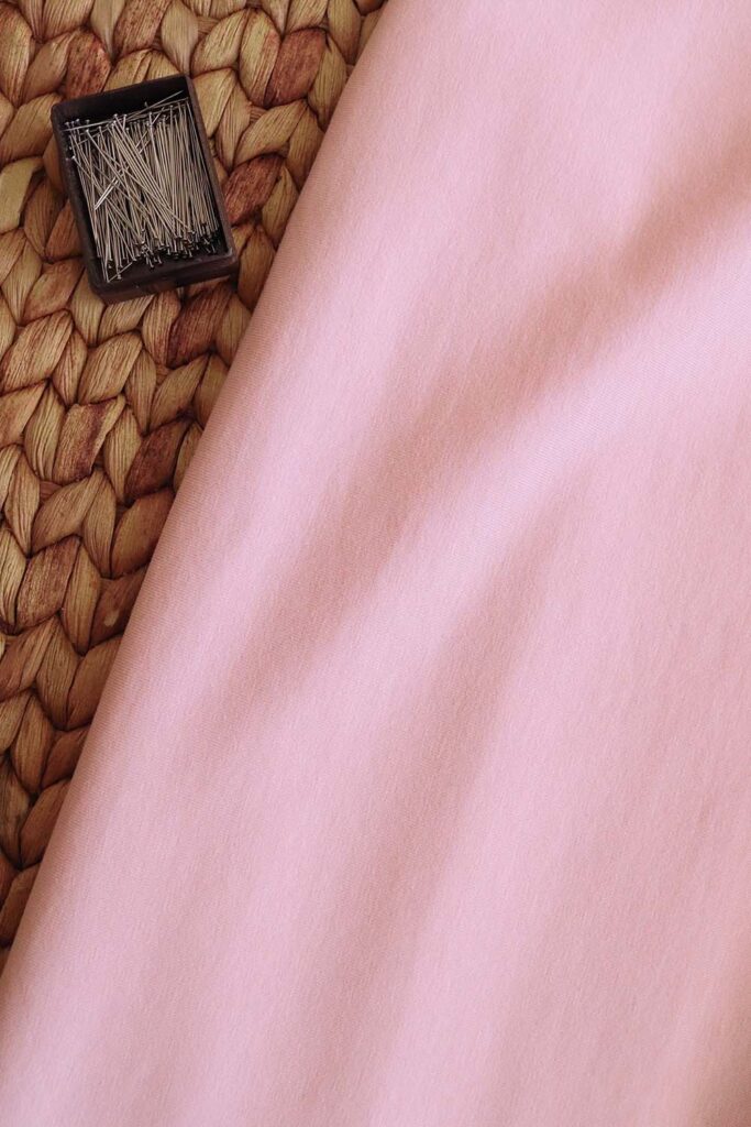 Tela de punto jersey de algodón orgánico de color rosa palo. Al lado se encuentra una pequeña caja de alfileres.