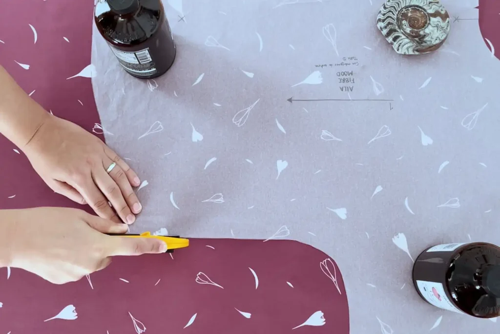 Una mano corta con cúter rotatorio de color amarillo una tela de color ciruela con motivos de hojas y flores blancas. La otra mano sujeta un patrón de una blusa, que a su vez, también está sujetado con botellas de vidrio a modo de pesas.